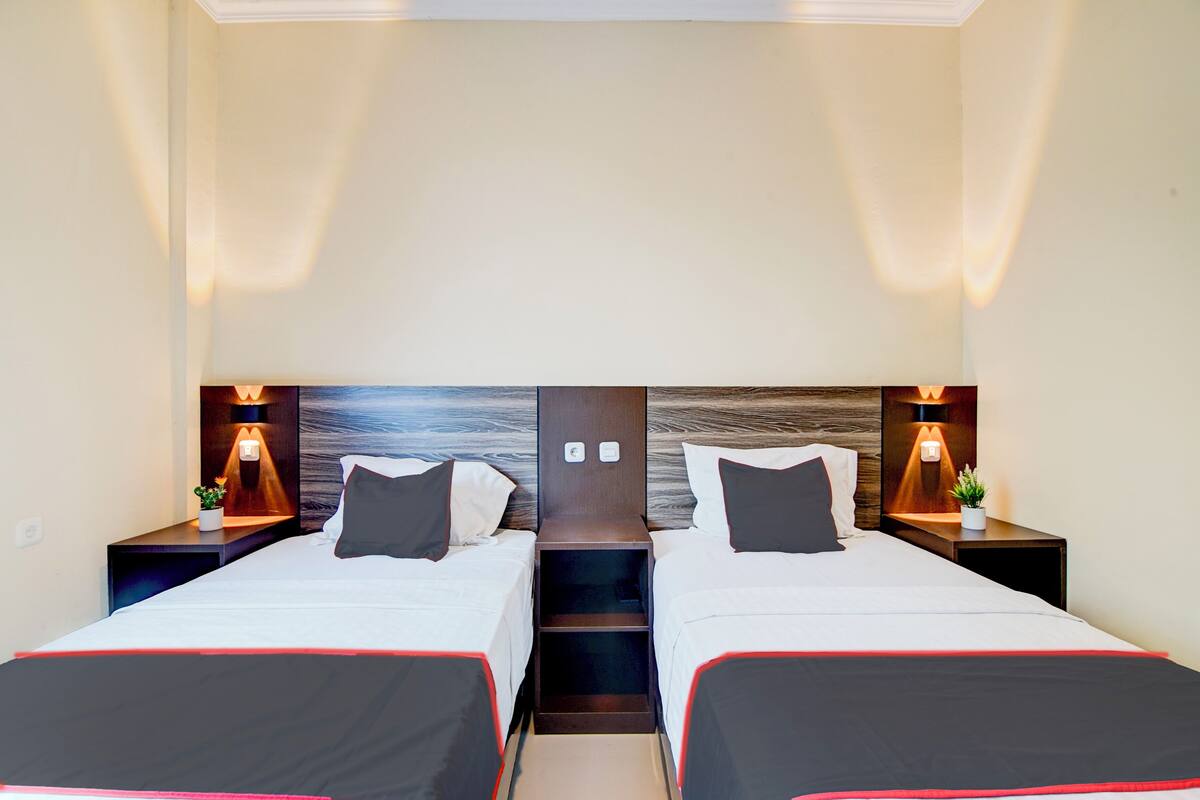 Habitación de hotel con dos camas, cada una con cabecero de madera, lámparas de noche y una planta pequeña en la mesita.