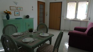 A dining area with a table set for four, surrounded by chairs. There is a green cabinet with decorative items on top, a door, and a window with curtains.
