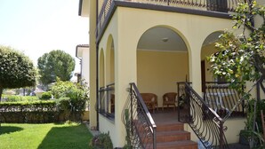 A yellow building with a staircase leading to an entrance, surrounded by greenery and a small patio area with chairs.