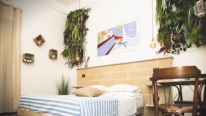 Un dormitorio con cama de madera, cabecera de ladrillo, una mesita y sillas. Hay plantas en macetas en la pared y un cartel sobre la cama.