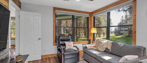 Een woonkamer met een open haard, een bank, een fauteuil en een groot raam met uitzicht op de omgeving.