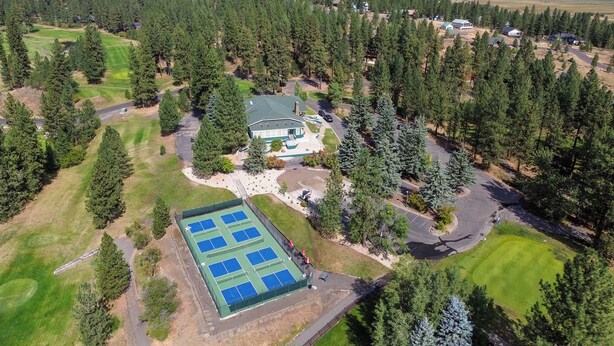 An aerial view of a property with a large tennis court, surrounded by dense forest, and a main building with a green roof.