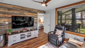 A modern living room with a large flat-screen TV, a ceiling fan, wooden wall paneling, a black leather sofa, and a large window offering a view of the outdoors.