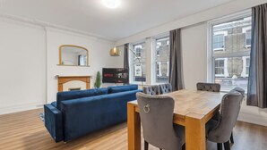 A modern living room with a blue sectional sofa, a wooden dining table with four chairs, a flat-screen TV, and large windows with curtains. - Modern 5-Bedroom Fulham Flat - Sleeps 12 (London)