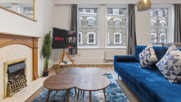 A living room with a blue sofa, a fireplace, a TV on a stand, and a view of buildings through large windows. - Modern 5-Bedroom Fulham Flat - Sleeps 12 (London)