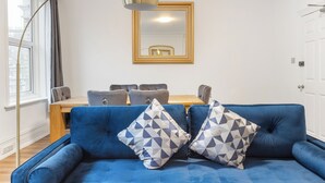 A room with a blue sofa, two patterned pillows, a wooden table with chairs, and a large mirror on the wall. - Modern 5-Bedroom Fulham Flat - Sleeps 12 (London)