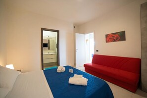A hotel room with a bed, a red sofa, a painting, and a bathroom visible through an open door.