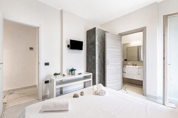 A modern bedroom with a large bed, a bedside table, a flat-screen TV mounted on the wall, and a bathroom visible through an open door.