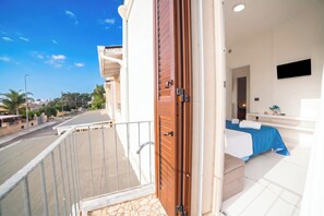 A balcony with a view of a street and buildings, a wooden door, a bed with a blue bedspread, and a flat-screen TV mounted on the wall. - Pino 3 - 3 bedrooms Near The Sea, Nardò, Italy (Nardò)