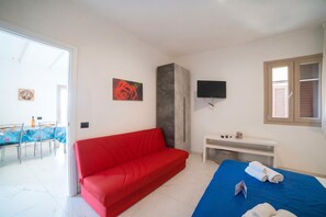A room with a red sofa, a bed with blue bedding, a TV mounted on the wall, a small table, and a window with blinds.