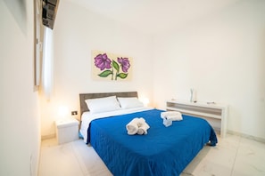 Une chambre d’hôtel avec un lit, deux tables de chevet blanches, une couvre-lit bleue et un tableau de fleurs accroché au mur.