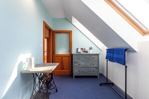 Ein kleiner Raum mit schrägem Dach, einer Holztür, einem Tisch mit Nähmaschine, einer Kommode und einem blauen Handtuch auf einem Ständer.