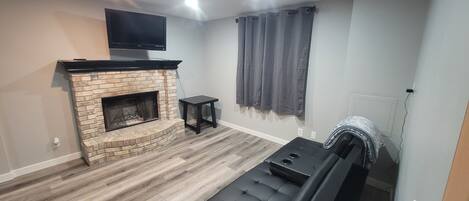 A modern living room with a fireplace, black leather sofa, and a flat-screen TV mounted on the wall.