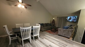 Ein Essbereich mit Tisch und Stühlen, ein Wohnzimmer mit Sofa und Fernseher, ein Weihnachtsbaum und ein Deckenventilator.