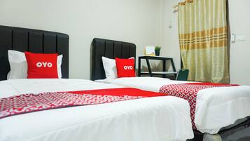 A hotel room with two beds, each with a black headboard and white bedding. There are red pillows with the word "OYO" on them. A small table with a plant and a calendar is visible in the corner.