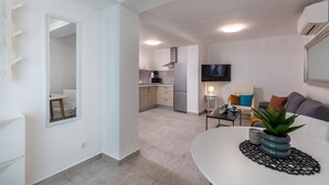 Una sala de estar moderna con un sofá blanco, una mesa de centro y un televisor. Se observa una cocina al fondo con un horno microondas y una estufa.