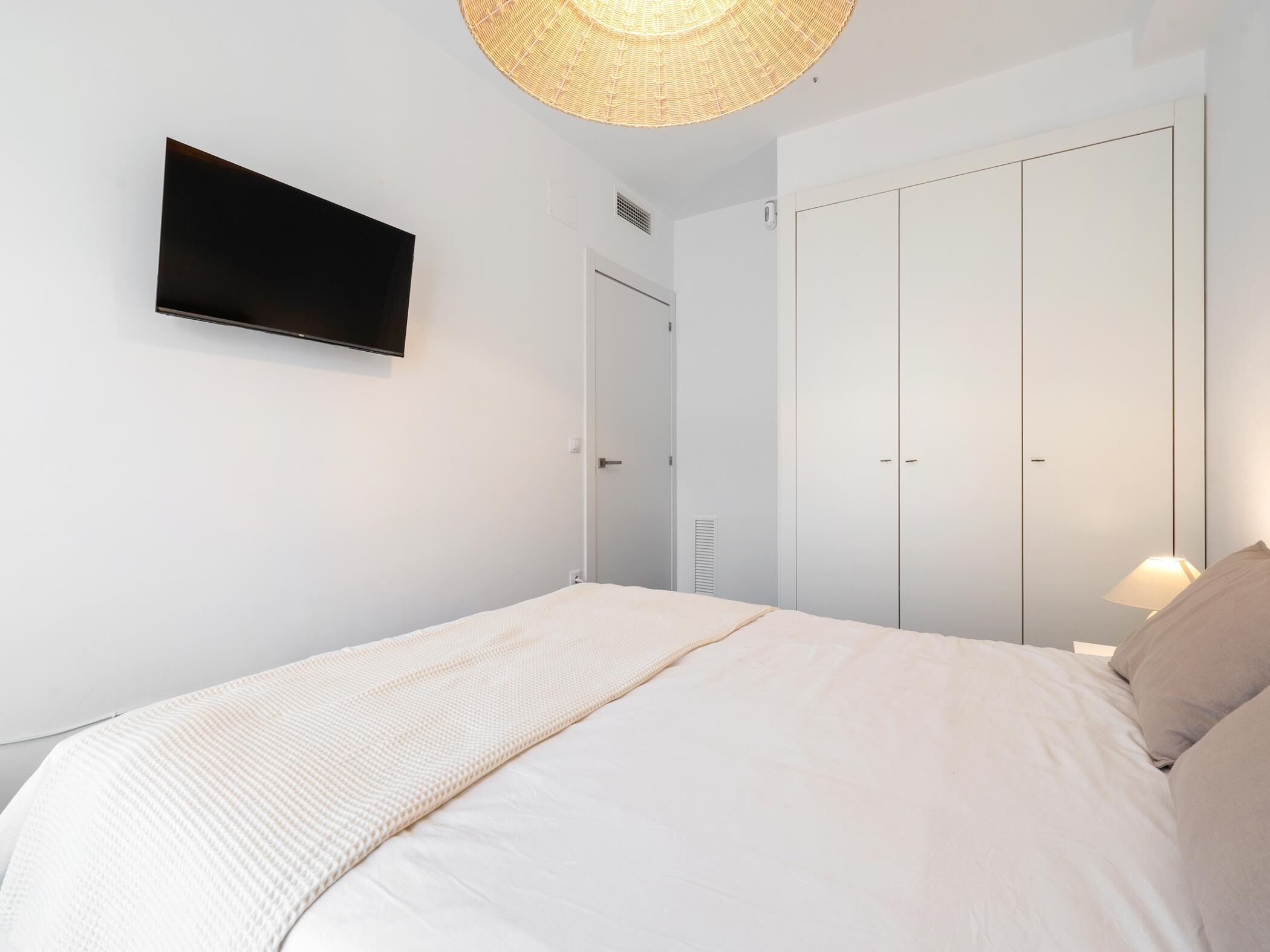 Une chambre à coucher avec un lit, un téléviseur fixé au mur, une armoire et une lampe.