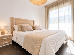 Ein Schlafzimmer mit Holz-Kopfteil, einem Nachttisch mit Lampe, einem großen Bett mit beiger Decke und Blick ins Freie durch leichte Vorhänge.