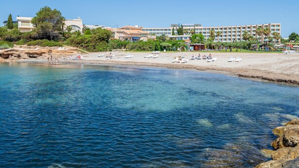 Un complejo turístico en la playa con aguas azules cristalinas, tumbonas y un gran edificio de hotel al fondo.