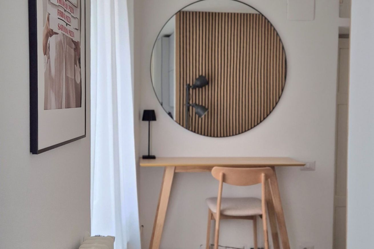 A room with a round mirror, a wooden desk, a chair, and a framed picture on the wall.