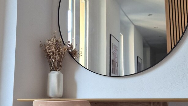 Een ronde spiegel aan een muur, een houten stoel en een witte vaas met gedroogde bloemen.