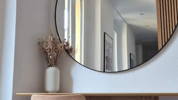 Een ronde spiegel aan een muur, een houten stoel en een witte vaas met gedroogde bloemen.