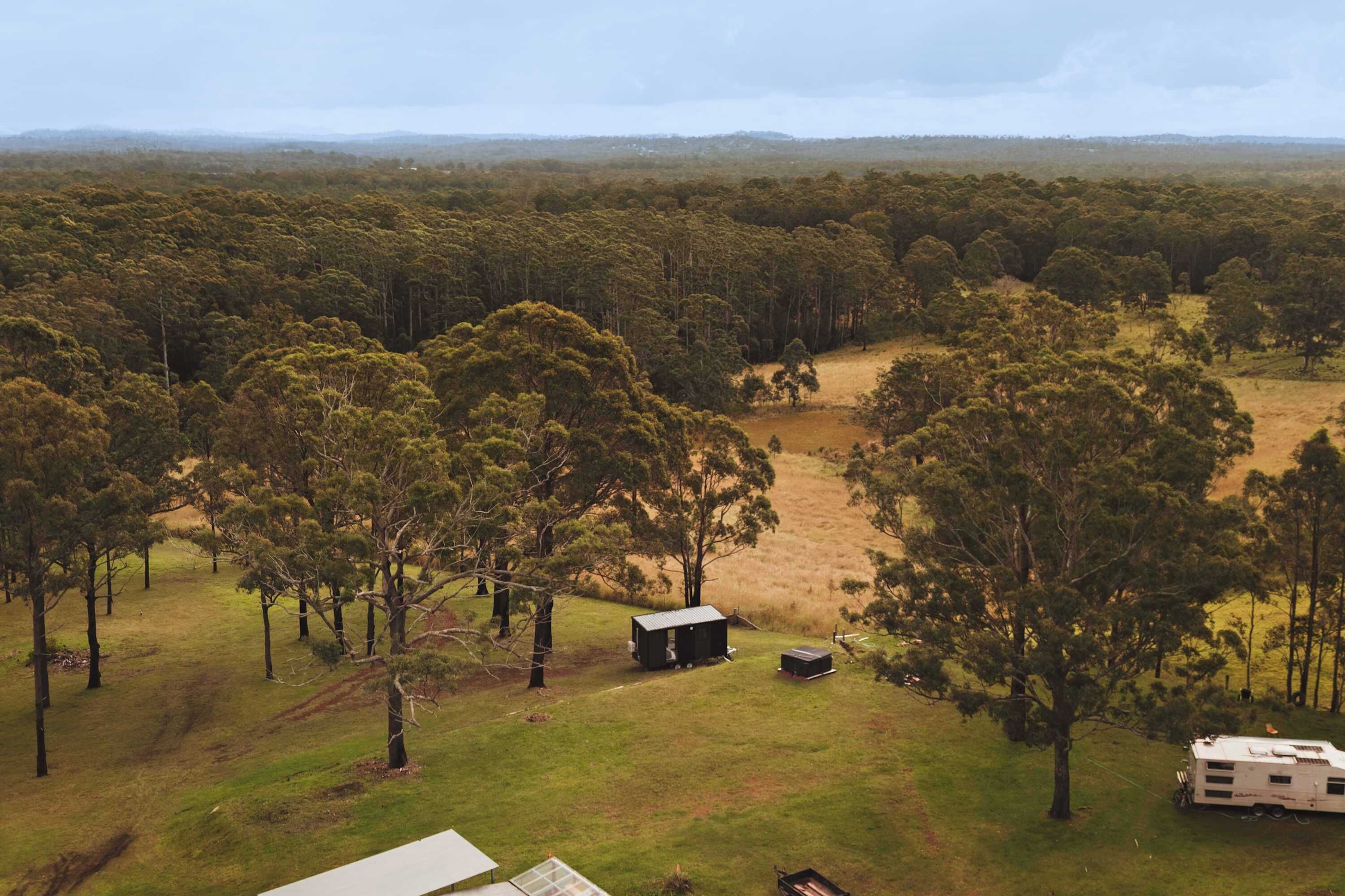 Un paysage rural avec une petite cabane et une caravane, situés dans une zone herbeuse entourée d’arbres.