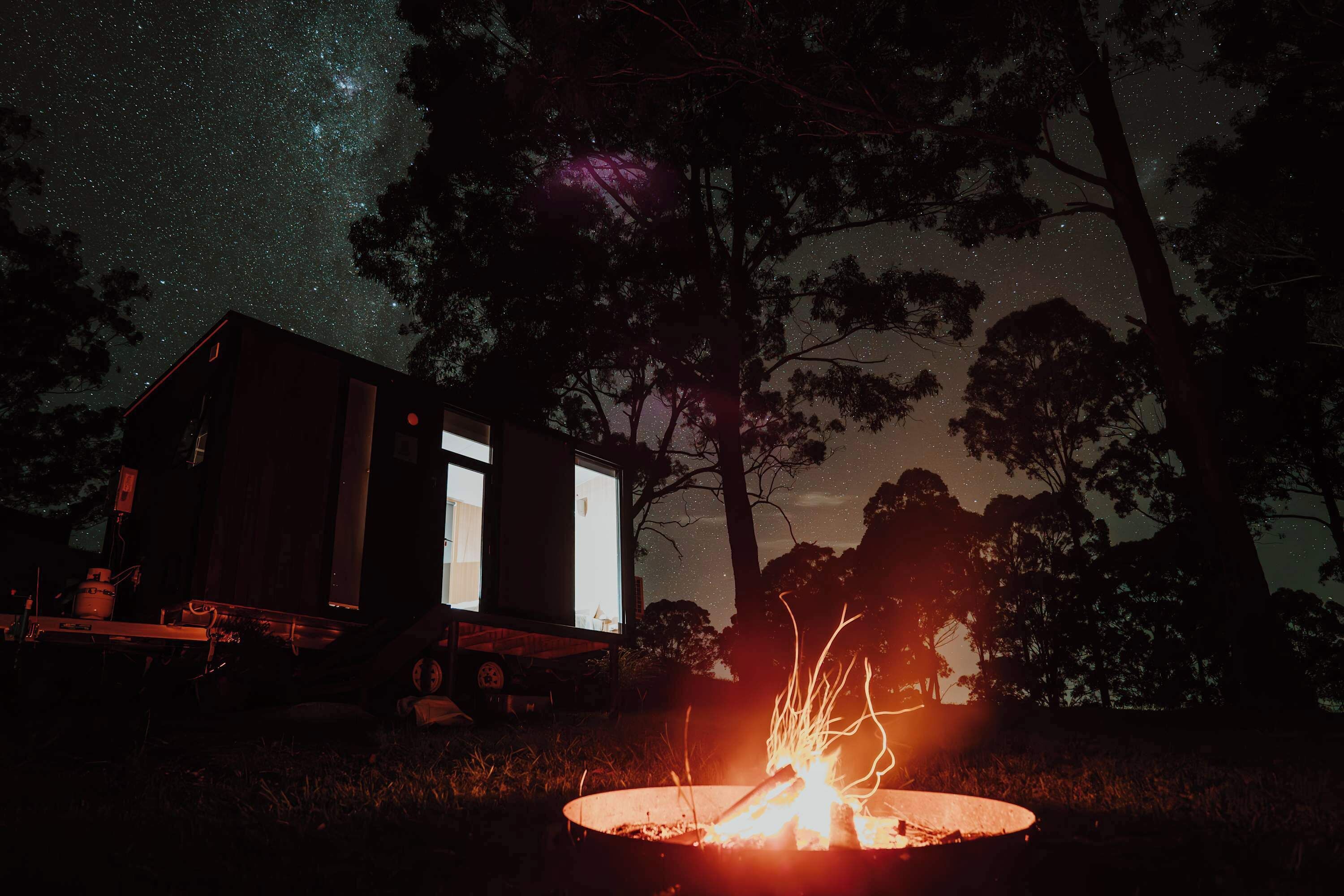 A cabin with a fire pit in front, surrounded by trees under a starry night sky.