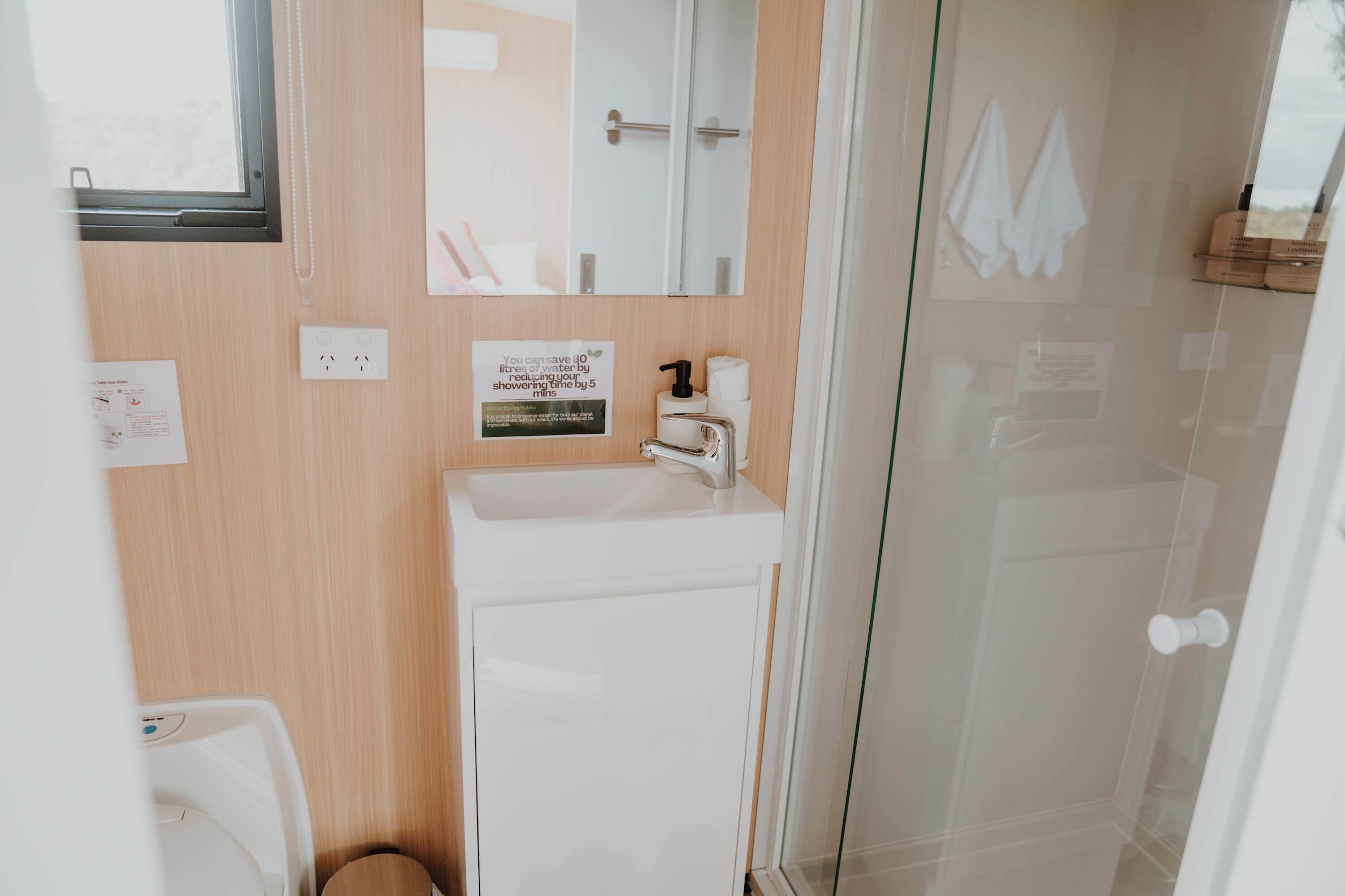 A bathroom with a sink, mirror, and shower. There are toiletries and a sign about water conservation.