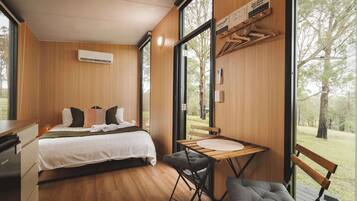 A modern bedroom with a bed, bedside table, and a desk with a chair. There is a large window providing a view of the outdoors.