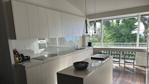 A modern kitchen with white cabinetry, a large island, and a view of greenery outside. - Luxury Beach House on two acres / Private Bathing Box / Botanic Like Gardens (Dromana)