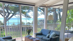 A modern living room with a large glass door leading to a balcony with a view of the ocean. There is a blue sofa, a beige sofa, and a wooden coffee table with books. - Luxury Beach House on two acres / Private Bathing Box / Botanic Like Gardens (Dromana)