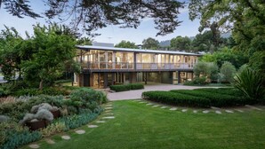A modern two-story house with a large glass facade, surrounded by a well-maintained garden with various plants and a stone pathway leading to the entrance. - Luxury Beach House on two acres / Private Bathing Box / Botanic Like Gardens (Dromana)