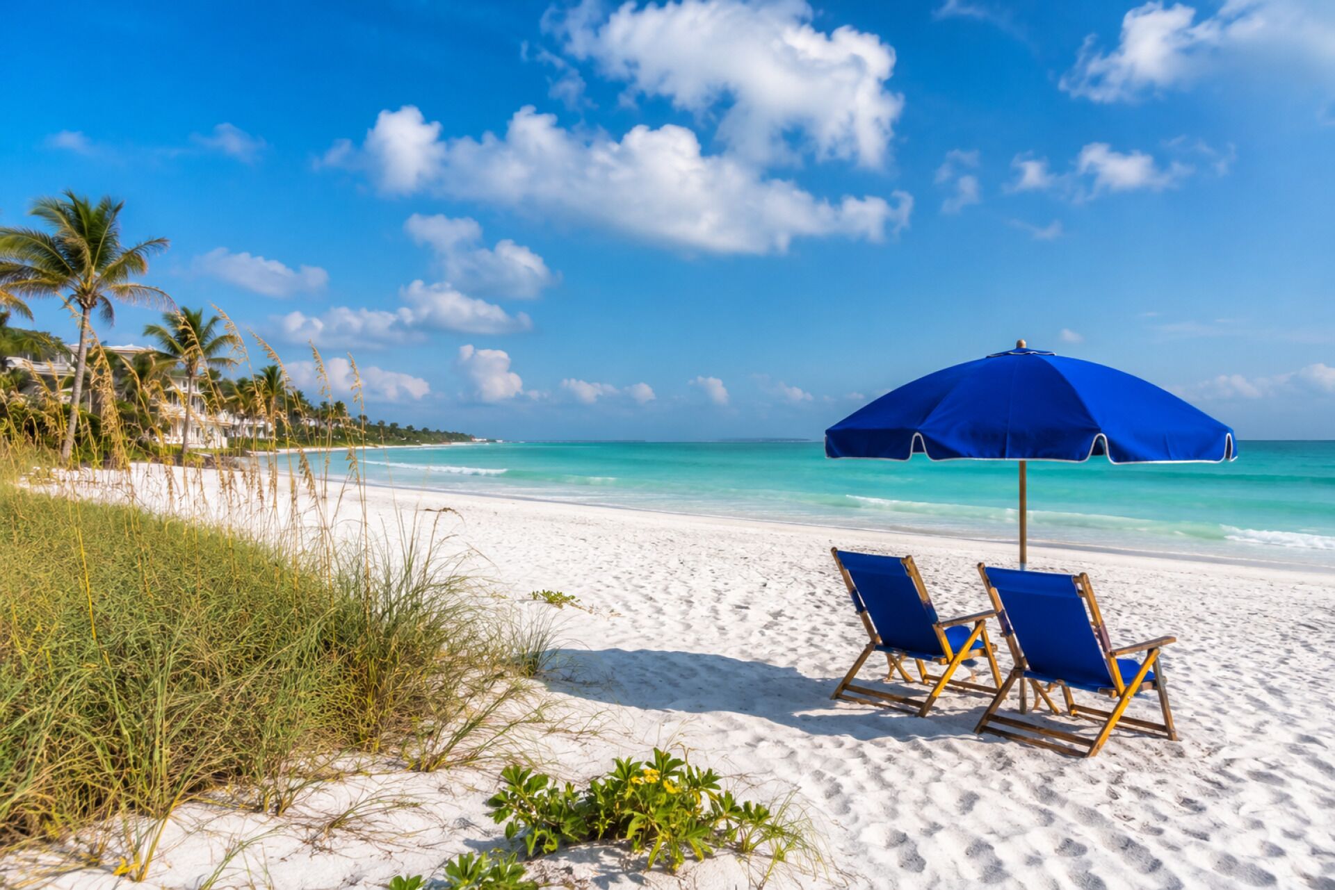 A beach with white sand, a blue umbrella, and two blue lounge chairs.