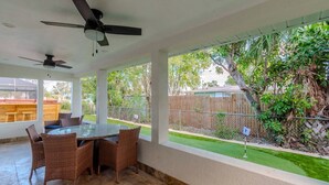 Une terrasse couverte avec une table et des chaises, un ventilateur de plafond et une vue sur une cour arrière clôturée, agrémentée de verdure.