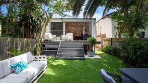 Una casa moderna con un patio trasero que incluye un sofá blanco, una mesa de comedor y una escalera que lleva a una terraza.