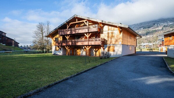A wooden chalet with a balcony, a paved driveway, and a mountainous backdrop. - Beautiful, bright apartment for 6 pers (Morillon)