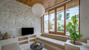 Una sala de estar moderna con pared de piedra, un televisor de pantalla plana, un sofá color beige, una mesa de centro y ventanas amplias con vistas a follaje.