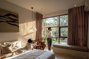Un dormitorio con un ventanal grande, una cama, un sillón de lectura y una persona de pie junto a la ventana.