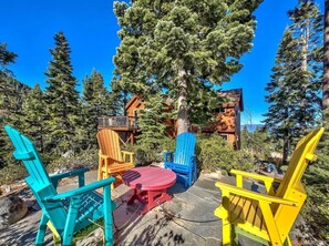 Eine Terrasse mit bunten Adirondack-Stühlen und einem roten Tisch, umgeben von Bäumen und einer Holzhütte.