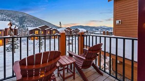 Une terrasse en bois avec des fauteuils Adirondack, une vue sur des chalets enneigés et un paysage montagneux en arrière-plan.