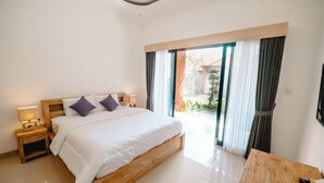 A bedroom with a wooden bed, nightstands, a wooden table, and a view of the outdoors through a sliding glass door. - Kubu Dangin Guest House Kerobokan  (Keroboken Kelod)