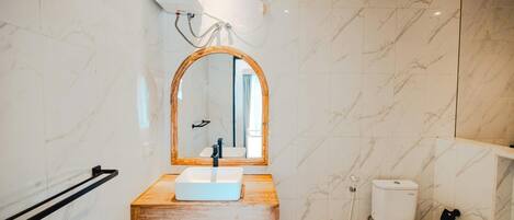 A bathroom with a wooden vanity, a white sink, a mirror with a wooden frame, a toilet, and marble walls.
