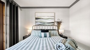 Ein ordentlich bezogenes Bett mit gestreifter Bettwäsche, eine Nachttischlampe und ein an der Wand hängendes Kunstwerk.