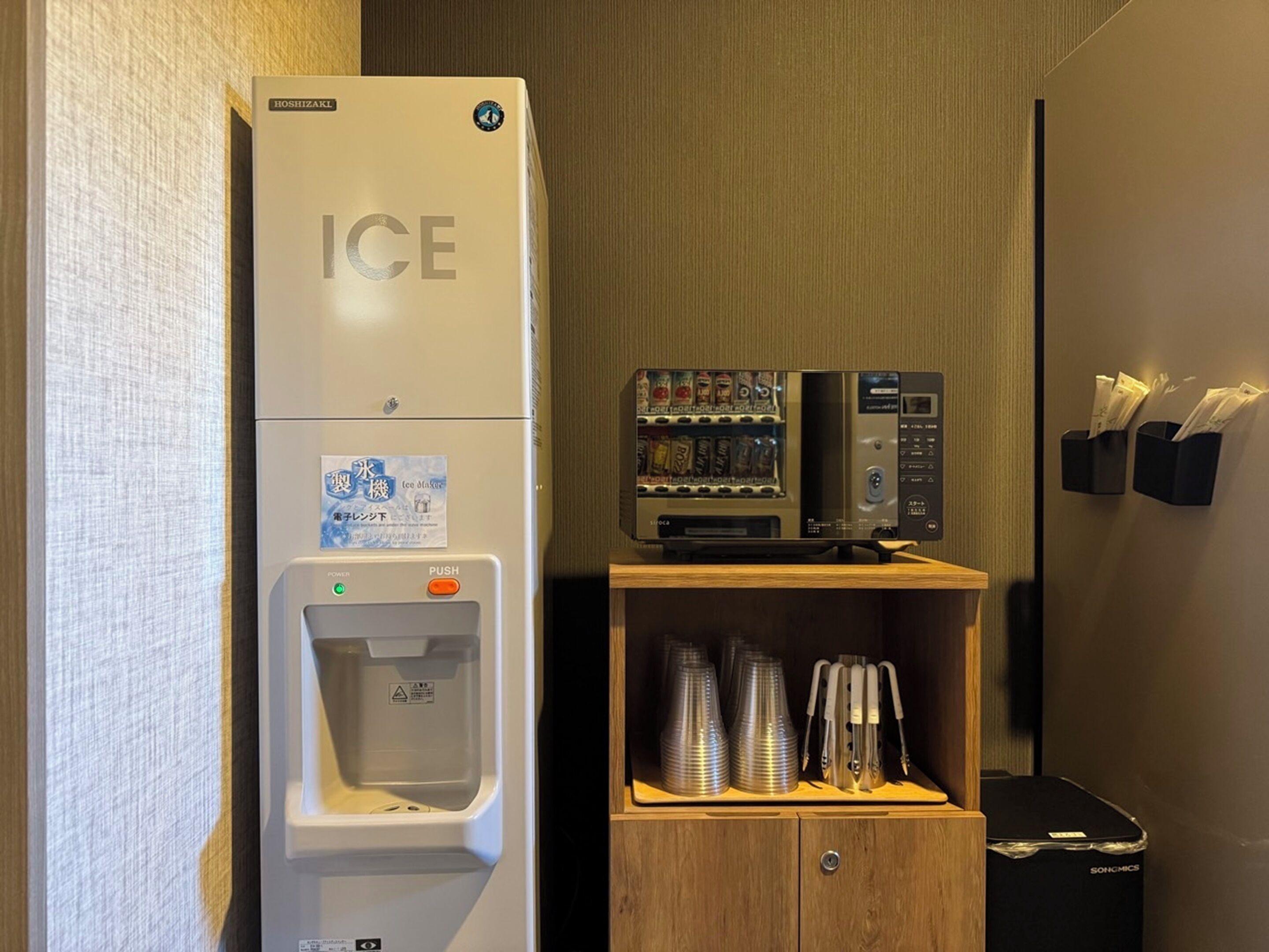 A vending machine with ice and beverages, a water dispenser, and a cabinet with stacked cups and a trash bin.