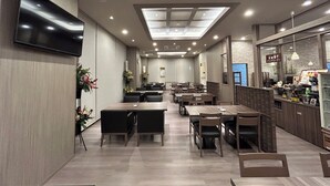 A modern restaurant with multiple dining tables and chairs, a flat-screen TV mounted on the wall, and a kitchen area visible through a glass partition.