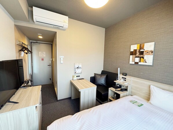 A hotel room with a bed, a desk, a chair, a TV, and an air conditioning unit.