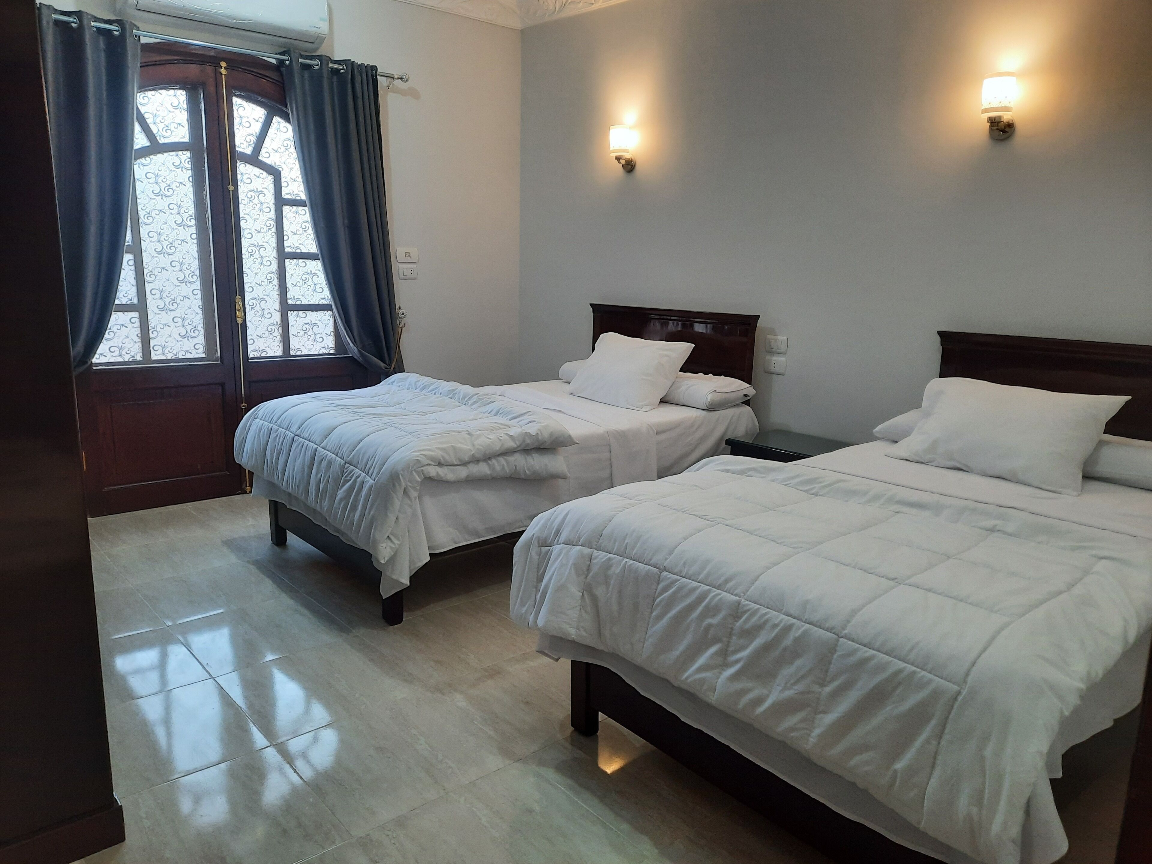 Quarto de hotel com duas camas, uma janela com cortinas e luminárias fixadas na parede.