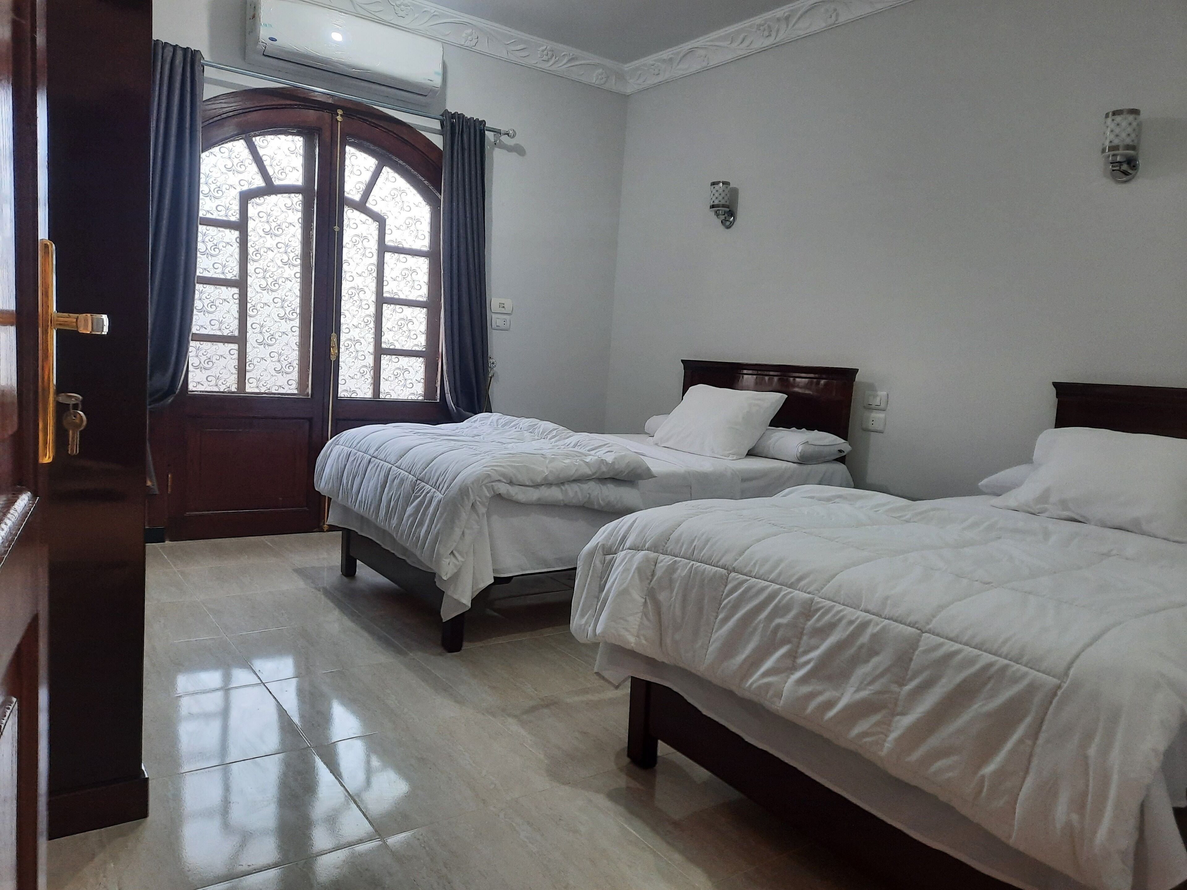 A hotel room with two beds, a wooden door, a window with curtains, and an air conditioning unit.
