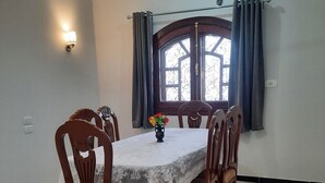 A dining area with a table set for six, surrounded by wooden chairs. A decorative vase with flowers is on the table. The room has a tiled floor and a window with curtains. - Charming 2-bedroom apartment with WiFi, AC in wonderful Luxor Governorate . (Luxor Governorate)